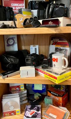 Wooden bookshelf filled with vintage electronics, including cameras, a camcorder, blank CD-Rs and DVD-RWs, alongside books, a novelty...