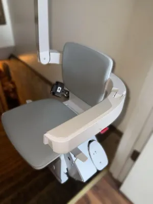 Modern motorized stair lift chair with grey vinyl padding and a white plastic frame, shown installed on a staircase.