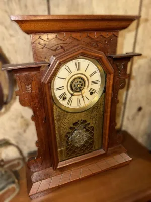 Antique gingerbread mantel clock in a carved walnut case, featuring a Roman numeral dial and an ornate gold-tone pendulum window.