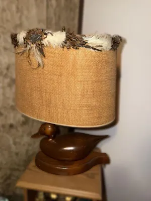 Vintage carved wooden duck decoy table lamp, featuring a burlap shade embellished with a decorative feather trim.