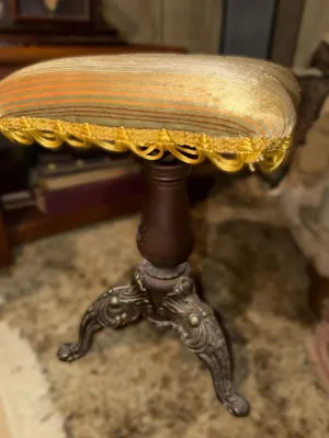 Antique piano stool featuring an upholstered striped seat with yellow fringe trim, supported by a turned wood pedestal and ornate cast...