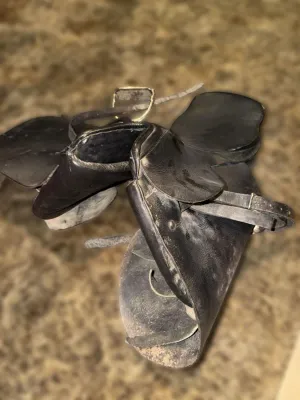 Vintage black leather horse tack, including stirrup leathers and flaps, showing signs of heavy wear and dust.