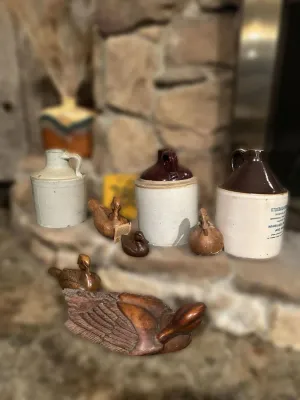 Collection of vintage stoneware whiskey jugs and hand-carved wooden duck decoys of various sizes, displayed on a stone hearth.