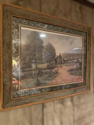 Framed rustic landscape print depicting a country cabin and dirt road, housed in a weathered wood frame with rope trim detailing.