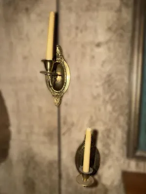 Pair of vintage brass wall sconces with oval backplates and decorative scrollwork, holding cream-colored taper candles.