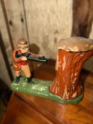 Vintage cast iron mechanical bank featuring Teddy Roosevelt aiming a rifle at a tree stump, showing some paint wear consistent with age.