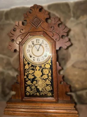 Antique gingerbread mantel clock featuring an ornate carved oak case and a gold-stenciled glass door depicting floral and urn motifs.