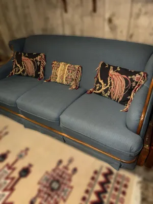 Vintage blue upholstered three-cushion sofa with wood trim, styled with three decorative fringed throw pillows featuring bird and floral...