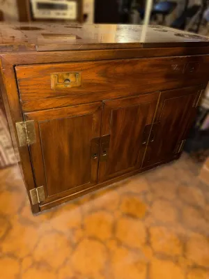 Vintage campaign-style wooden bar cabinet featuring brass flush-mount hardware, a top drawer, and lower cabinet doors.