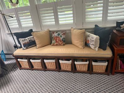 Long wooden storage bench with a tan upholstered cushion, six woven wicker storage baskets, and an assortment of autumn-themed throw...