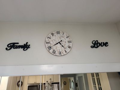 Rustic farmhouse-style wall clock with a distressed white wood finish and Roman numerals, flanked by black cursive wall signs reading...