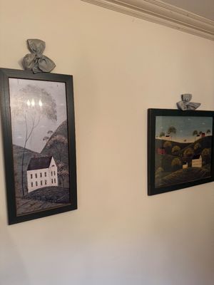 Pair of framed folk art prints depicting rural landscapes with houses and rolling hills, presented in dark frames with decorative fabric...