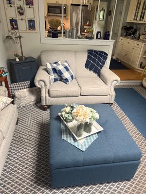 Contemporary beige upholstered loveseat with rolled arms and nailhead trim, paired with a large blue fabric storage ottoman used as a...