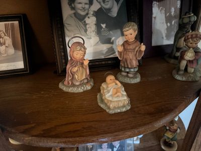 Goebel Hummel nativity figurines, including Mary, Joseph, and Baby Jesus, displayed on a wooden shelf alongside vintage framed photographs.