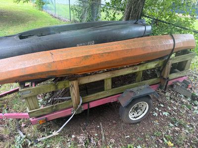 Two kayaks, including a dark green Old Town model and an orange model, strapped to a small utility trailer with wooden side rails.