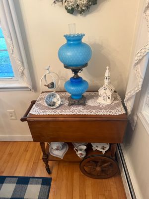 Vintage wooden drop-leaf tea cart featuring a blue hobnail glass hurricane lamp, a ceramic church figurine, and decorative tea sets on...