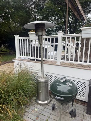 Stainless steel outdoor patio heater alongside a green Weber charcoal kettle grill, both showing light outdoor wear.