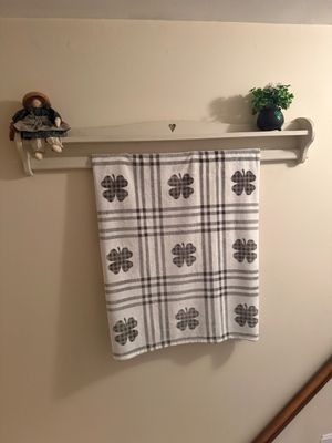 Country-style white wooden wall shelf with a heart cutout, featuring a hanging plaid shamrock towel, a small rag doll, and a potted faux...