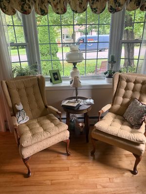 Pair of vintage tufted wingback armchairs in a gold fabric, flanking a round wooden side table with cabriole legs.