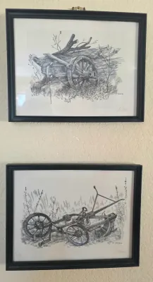 Pair of framed black and white ink drawings by J. Ader, depicting rustic farm equipment including a wagon wheel and a plow, presented in...