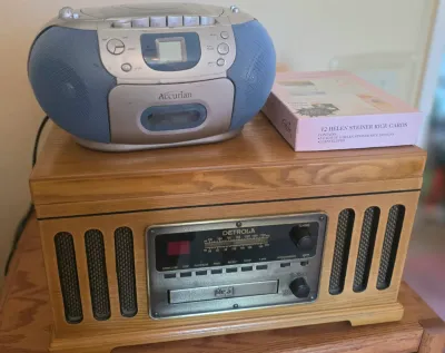 Detrola retro-style wooden music center with CD player and radio, topped with a blue Accurian portable CD/cassette boombox and a box of...