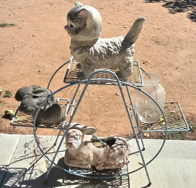 Vintage white wire ferris wheel plant stand holding a ceramic cat figurine, two small rabbit figurines, a deer planter, and a clear glass...