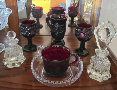 Collection of vintage glassware including ruby red pressed glass goblets, a mug, and a teacup with a clear beaded saucer, flanked by two...