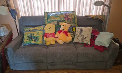 Grey upholstered three-cushion sofa styled with Winnie the Pooh plush toys, John Deere themed pillows, and miscellaneous textiles.