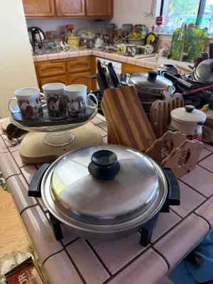 Vintage electric skillet with stainless steel lid, displayed alongside a wooden knife block, decorative mugs, and other kitchenware on a...