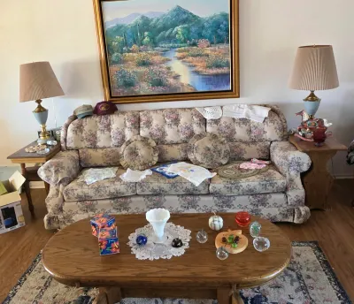Vintage floral upholstered sofa with matching round throw pillows, flanked by wooden end tables with brass and ceramic lamps, and an oval...