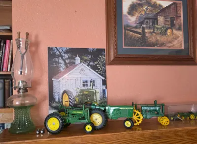 Collection of vintage John Deere die-cast toy tractors displayed on a wooden shelf alongside a green glass oil lamp and framed rustic...