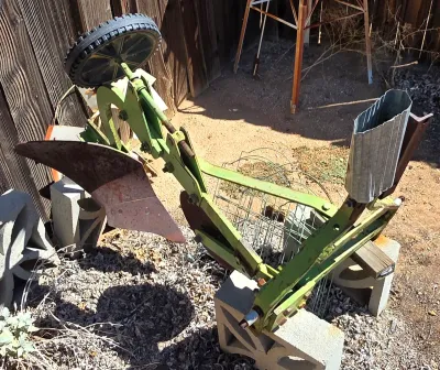 Vintage green walk-behind garden plow attachment with a rusty metal blade and a single rubber-treaded wheel, resting on concrete blocks.