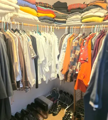 A walk-in closet filled with assorted clothing, including folded sweaters on top shelves, hanging shirts and jackets, and shoes arranged...