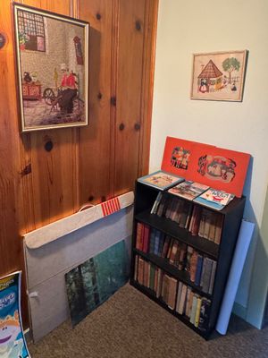 Small black three-shelf bookcase filled with vintage books, accompanied by framed needlepoint artwork and various prints.