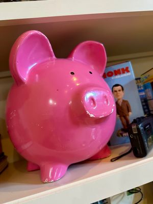 Large pink ceramic piggy bank with minor paint chipping on the snout and feet, displayed on a shelf alongside a Monk bobblehead and a...