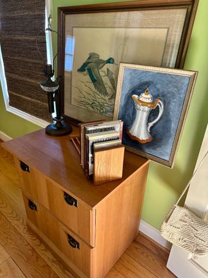 Two-drawer wooden filing cabinet or side table with black metal pulls, styled with framed artwork, a vintage lamp, and a wooden book rack.