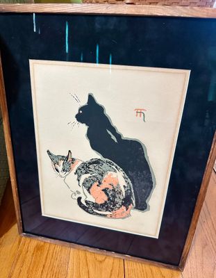 Framed Japanese woodblock print depicting a black cat and a calico cat, presented in a dark mat and a simple wooden frame.