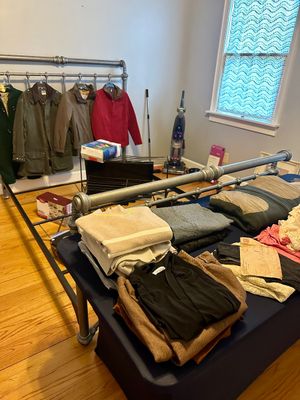 Industrial-style metal pipe bed frame displayed with various folded clothing items, jackets hanging on the headboard, and household goods...