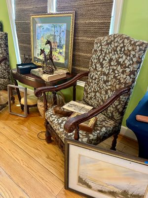 Traditional high-back armchair with leaf-patterned upholstery and carved wooden arms, shown alongside framed artwork, a small side table,...