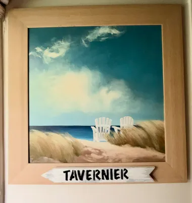 Framed beach landscape painting depicting two white Adirondack chairs on a sandy dune overlooking the ocean, featuring a decorative...