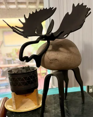 Rustic metal and river rock moose sculpture, featuring a natural stone body with black metal legs, head, and antlers.
