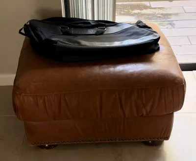 Vintage brown leather ottoman with nailhead trim along the base, topped with a black Harley-Davidson duffel bag.