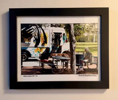 Framed color photograph titled Allan Cohen "AC" '19 by Stephanie Castro, depicting two people at a picnic table near a...