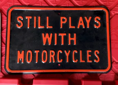 Embossed metal novelty sign reading "STILL PLAYS WITH MOTORCYCLES" in orange lettering on a black background.
