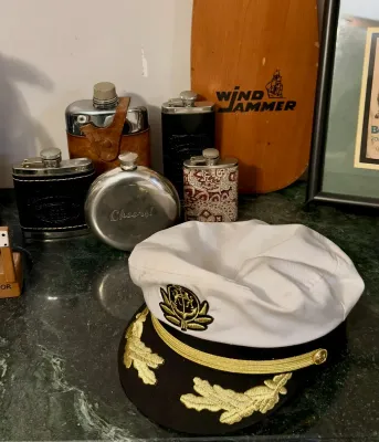 Nautical-themed collection featuring a white captain's hat with gold embroidery, several vintage metal flasks, and a wooden...