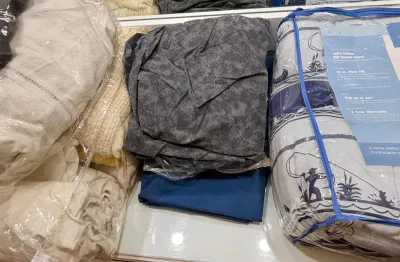 Assorted bedding items, including a packaged mattress pad, folded blue and grey sheets, and blankets in plastic storage bags.