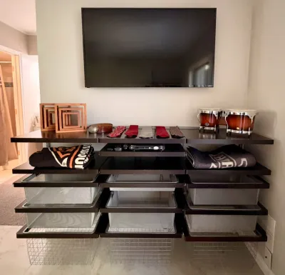 Modern dark wood wall-mounted shelving unit with integrated wire storage baskets, displaying various items including a flat-screen TV,...