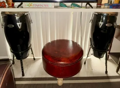 Pair of black Mano conga drums on metal stands, flanking a round faux-alligator leather ottoman with nailhead trim.