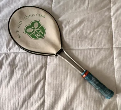 Vintage metal tennis racket with a blue wrapped grip, featuring a white zippered head cover printed with a green 'Bath and Tennis...