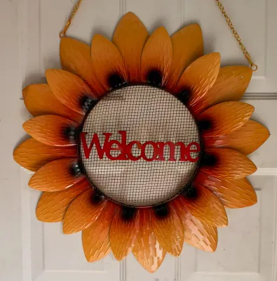 Decorative metal sunflower welcome sign featuring layered orange petals, a mesh center, and a hanging chain.